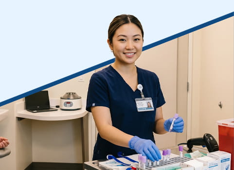 Phlebotomy technician wearing navy blue scrubs, a hospital name badge, and blue sterile gloves holding vials for collecting blood from patients in an exam room.