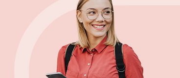 Young woman wearing glasses holding a smart phone in one hand and a travel cup of coffee in the other on a light pink background.