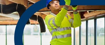 Man in yellow safety gear and blue hard hat installing duct.