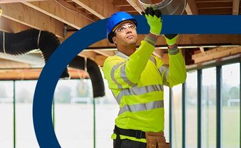 Man in yellow safety gear and blue hard hat installing duct.