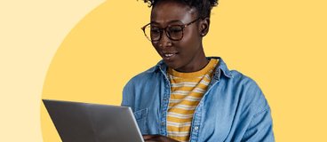 Woman wearing glasses standing in front of orange and pink background, looking at laptop she is holding.