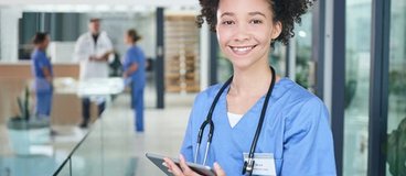 A woman with light skin and dark, curly hair wears scrubs and a stethoscope and holds a tablet