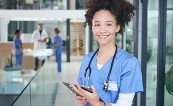 A woman with light skin and dark, curly hair wears scrubs and a stethoscope and holds a tablet