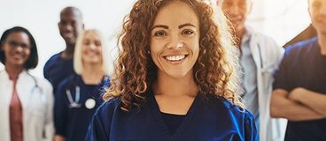 A woman with medium brown skin and curly brown hair wearing scrubs stands in the foreground; multiple people stand behind her wearing lab coats and scrubs and stethoscopes