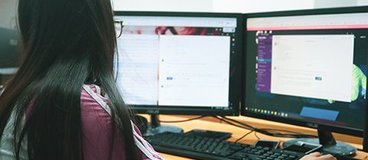 A woman with medium dark skin and dark hair watches two computer monitors