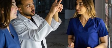 One person in a lab coat points at something, while two other people in scrubs look on