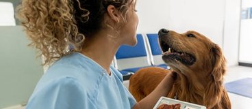 A woman with caramel skin and curly hair wears scrubs and holds an ipad while petting a dog