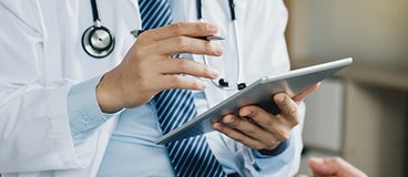 A person in a suit and tie wearing a lab coat and a stethoscope, holding an ipad and gesturing to another person with folded hands