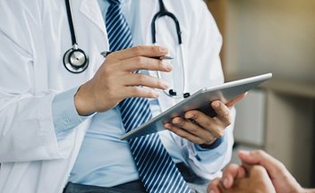A person in a suit and tie wearing a lab coat and a stethoscope, holding an ipad and gesturing to another person with folded hands