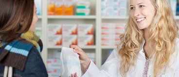 A woman with fair skin and blonde hair wears a lab coat and hands medication to a woman with medium skin and dark hair.