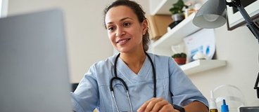 A woman wearing scrubs and a stethoscope and working on a laptop