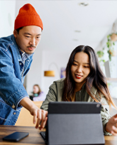 A woman with dark hair sits in front of a tablet, talking to a man in a red beanie who leans over her, pointing at the tablet