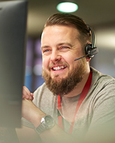 A man with light skin and brown hair, looking at a computer and talking on a headset while smiling