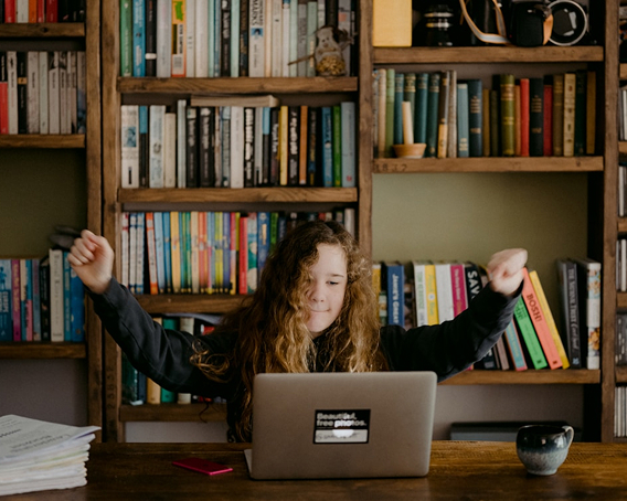 Teen girl in library with raised hands as she looks at laptop.
