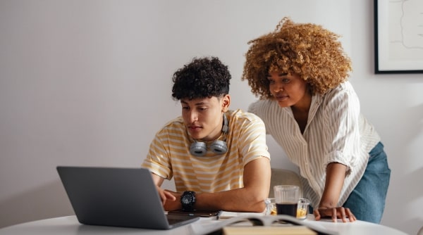 Mom helping teen boy using laptop at kitchen table.