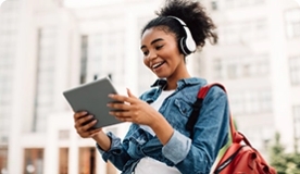 Teen girl with backpack and headphones smiling at tablet she's holding while outdoors.