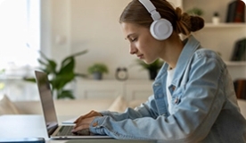 Teen girl wearing light denim jacket and white headphones studying on laptop in dining room.