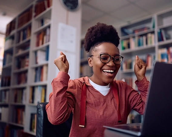 Teen girl wearing glasses and red striped hoodie celebrating something she sees on laptop in a library.