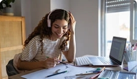 Teen girl in striped t-shirt wearing headphones taking notes as she studies on laptop computer.