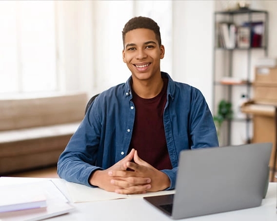 Young man wearing maroon t-shirt and denim button up smiling with hands folded at table in front of laptop.