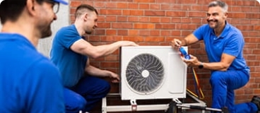 Three HVAC technicians inspecting and repairing an outdoor AC unit.