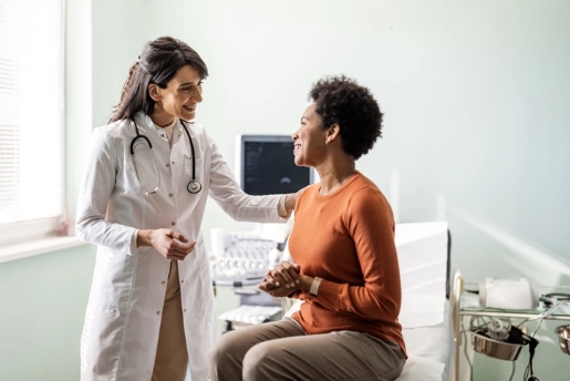 Healthcare worker in white coat with stethoscope speaking with a female patient in an exam room.