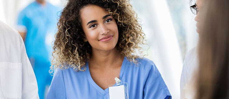 Woman in light blue scrubs with work badge clipped on collar smiling in group of healthcare professionals.