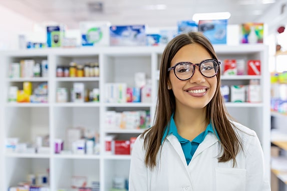 A pharmacy technician smiling in front of a shelf of medicine.