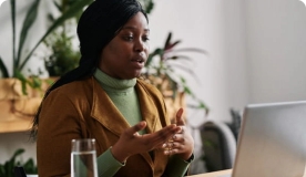 Woman in green sweater and brown jacket speaking to someone on laptop.
